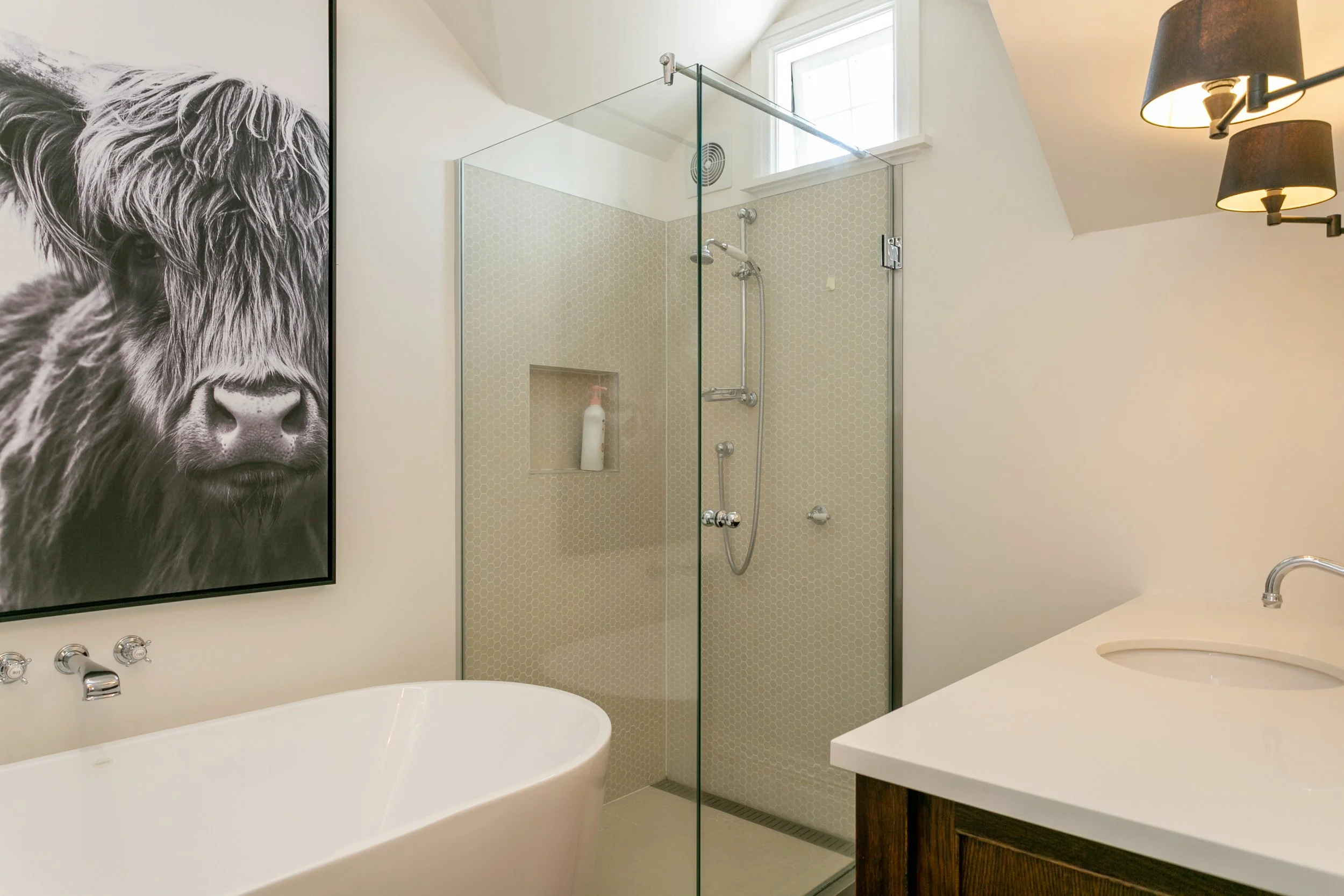 Modern bathroom with a black and white cow painting, a white bathtub, a glass shower with a built-in shelf, a small window, and a dark light fixture.