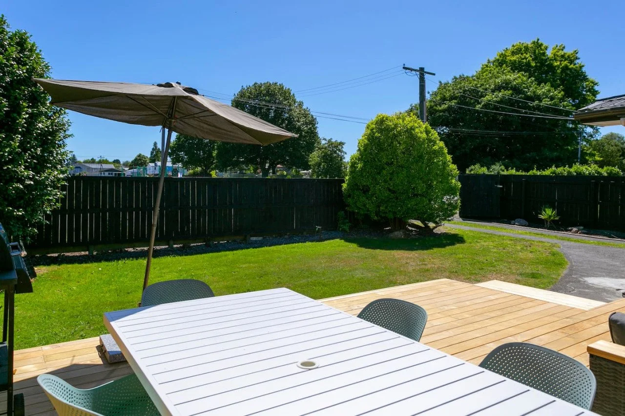 A backyard with a wooden deck, a white table, black chairs, a large umbrella, green grass, trees, a wooden fence, and a clear blue sky.