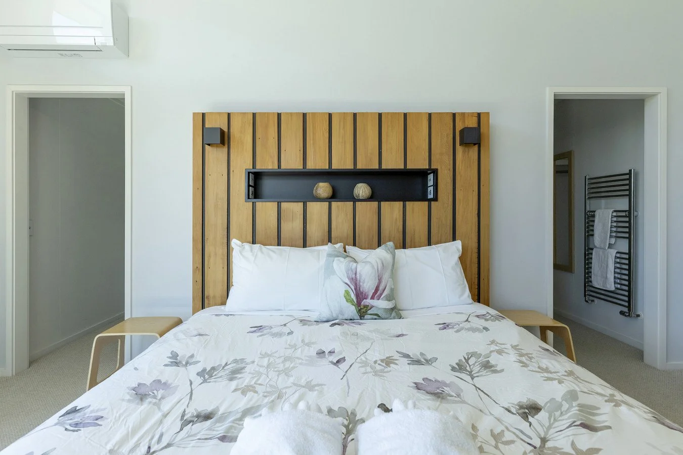 Bedroom with a bed featuring white bedding and pillows, a leaf pattern on the duvet, a wooden headboard with black accents, and small decorative objects on a shelf behind the bed.