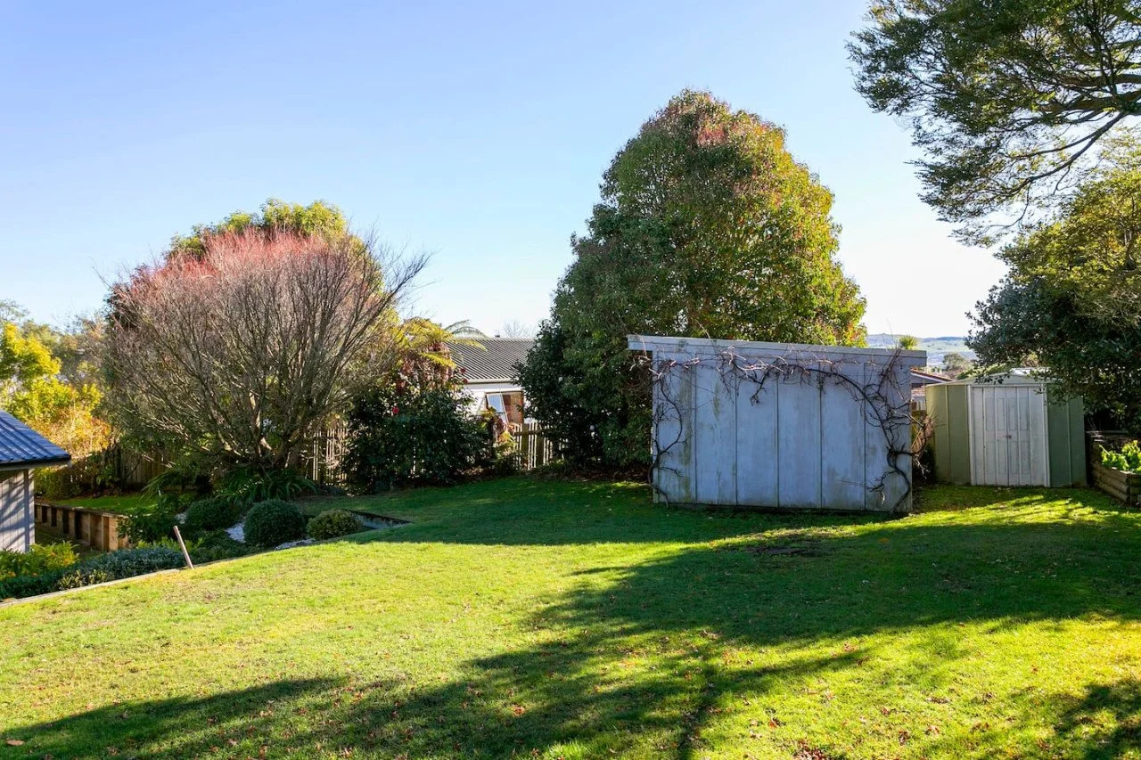 A backyard with green grass, trees, bushes, and two metal sheds, one gray and one green, with a house visible behind the trees under a clear blue sky.
