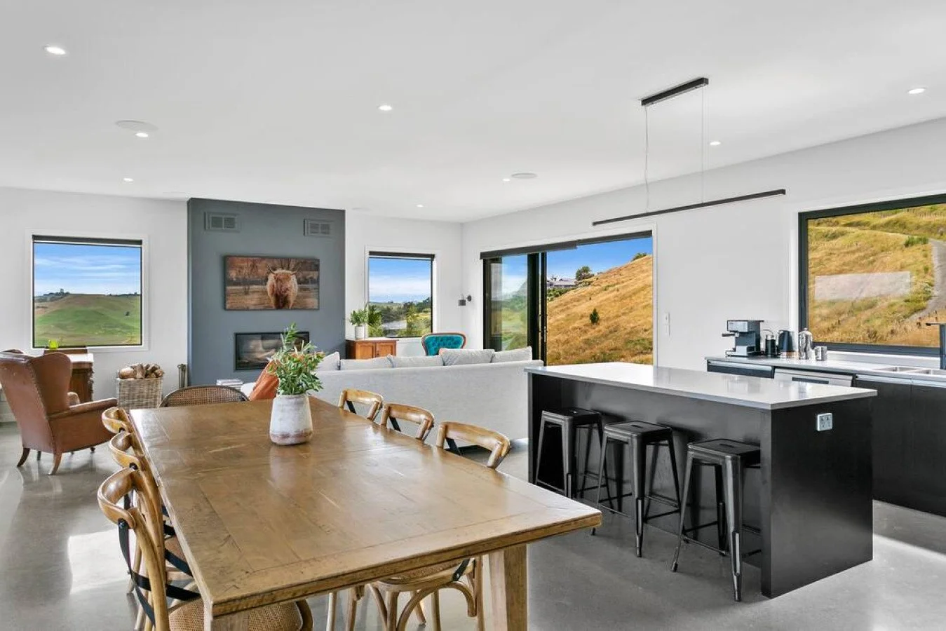 Open-concept living room and kitchen with large windows showing a hilly landscape outside, modern furniture, a wooden dining table with chairs, a gray accent wall with a painting, and a kitchen island with bar stools.