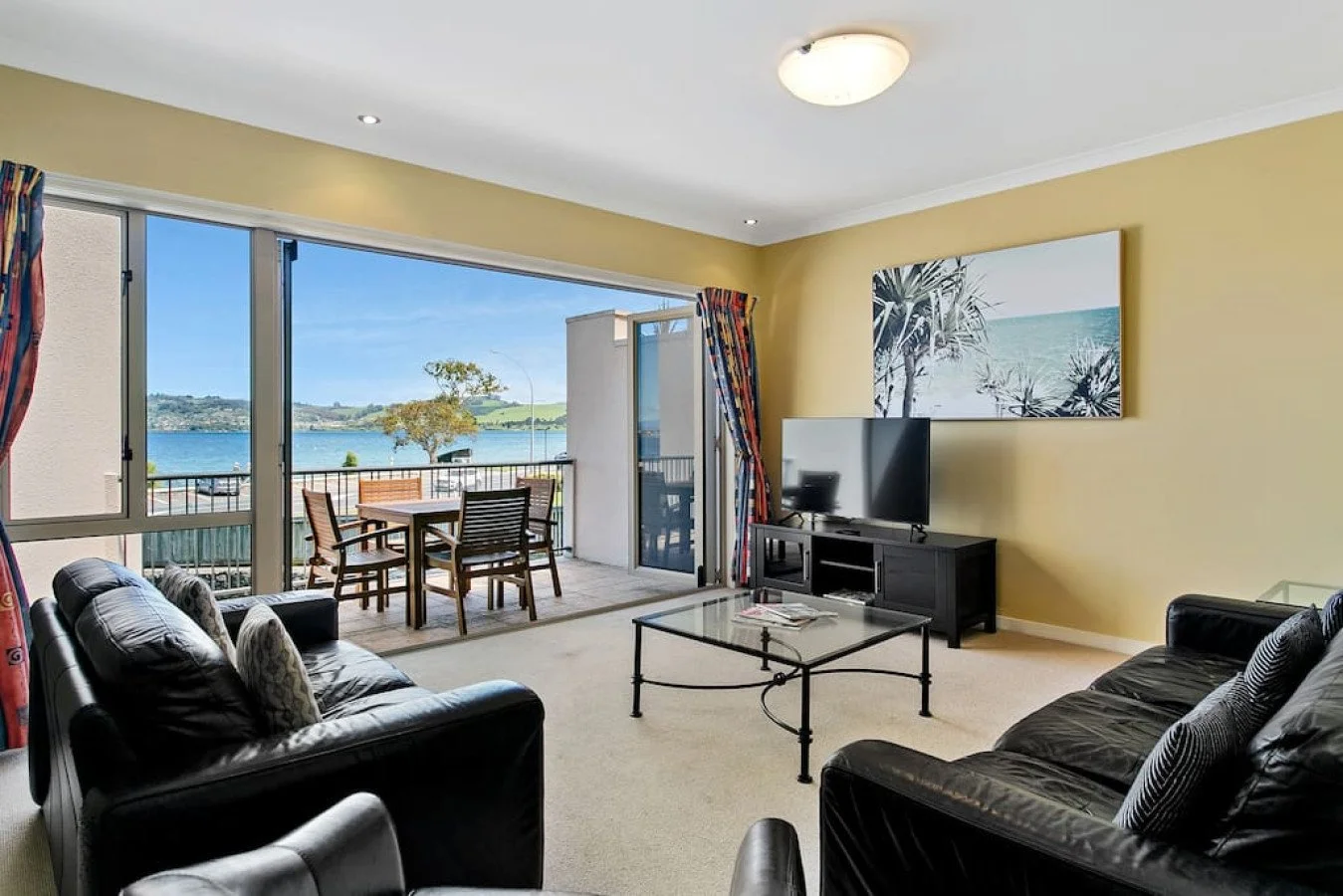 Living room with black leather sofas, a black TV stand with a flat-screen TV, a glass coffee table, and a large window and sliding door leading to a balcony with a lake view and outdoor dining furniture.