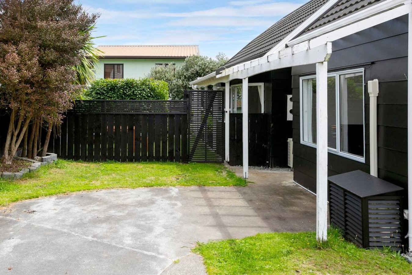 A backyard with a concrete patio, green grass, trees, a black wooden fence, and a black house with white trim and windows.
