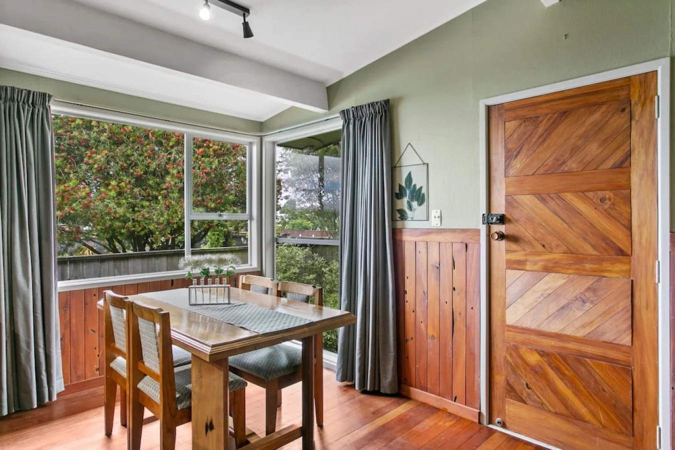 Dining area with wooden table and chairs, next to a large window with views of trees outside, and a wooden door with diagonal planks.