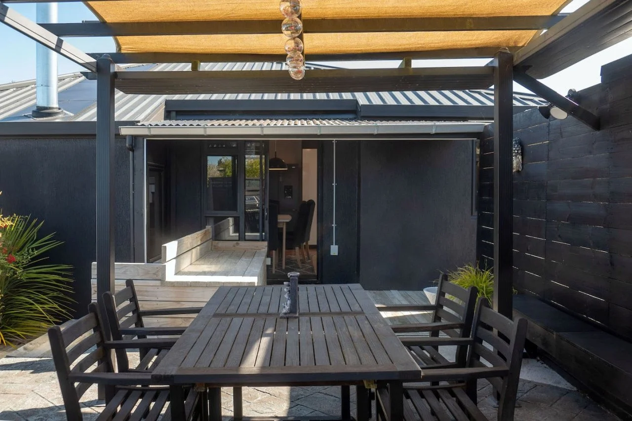 Outdoor patio with a wooden dining table and chairs, a garden area, and a black building with a sliding glass door leading to an indoor dining area.