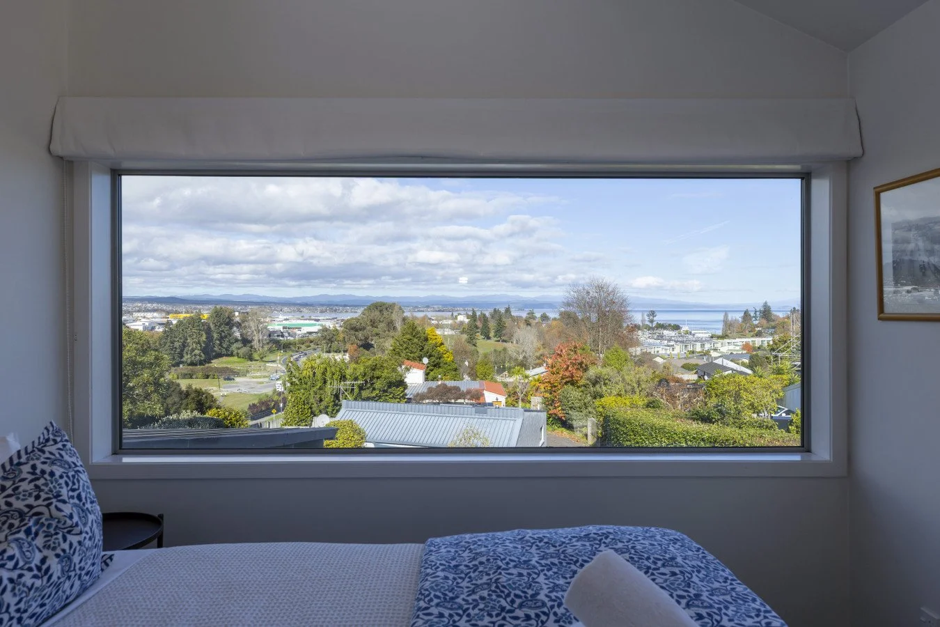 Room with a large window showing a view of trees, houses, and a distant body of water under a partly cloudy sky.