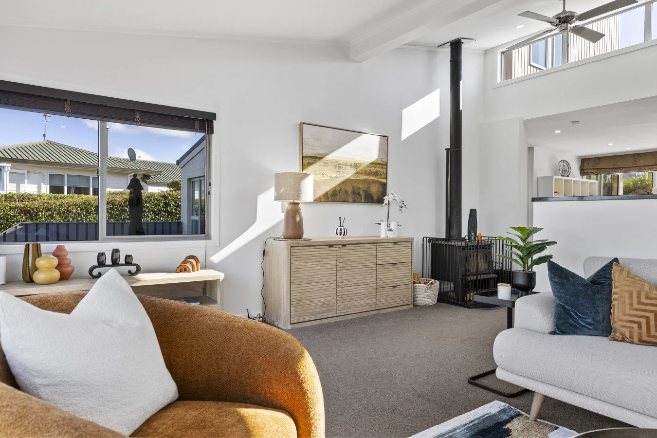 Living room with beige and white sofas, a wooden sideboard with a lamp and decorative items, a window with blinds, a black stove pipe, a potted plant, and a ceiling fan, illuminated by natural sunlight.