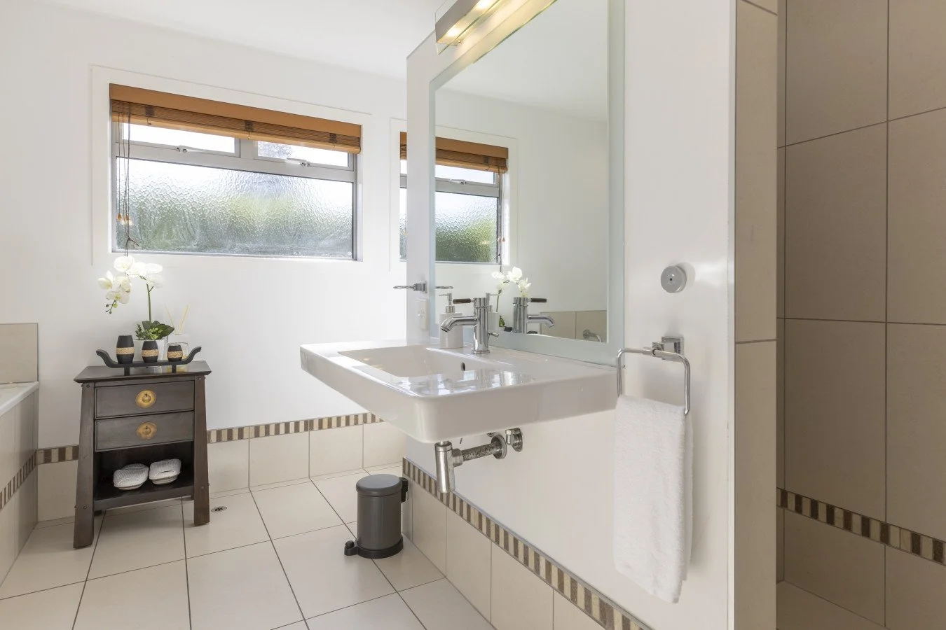 Modern bathroom with white walls, tiled floor, and large mirror over a white sink with two silver faucets. Two windows with frosted glass and brown blinds, decorative orchids on a dark side table, and a white towel hanging beside the sink.