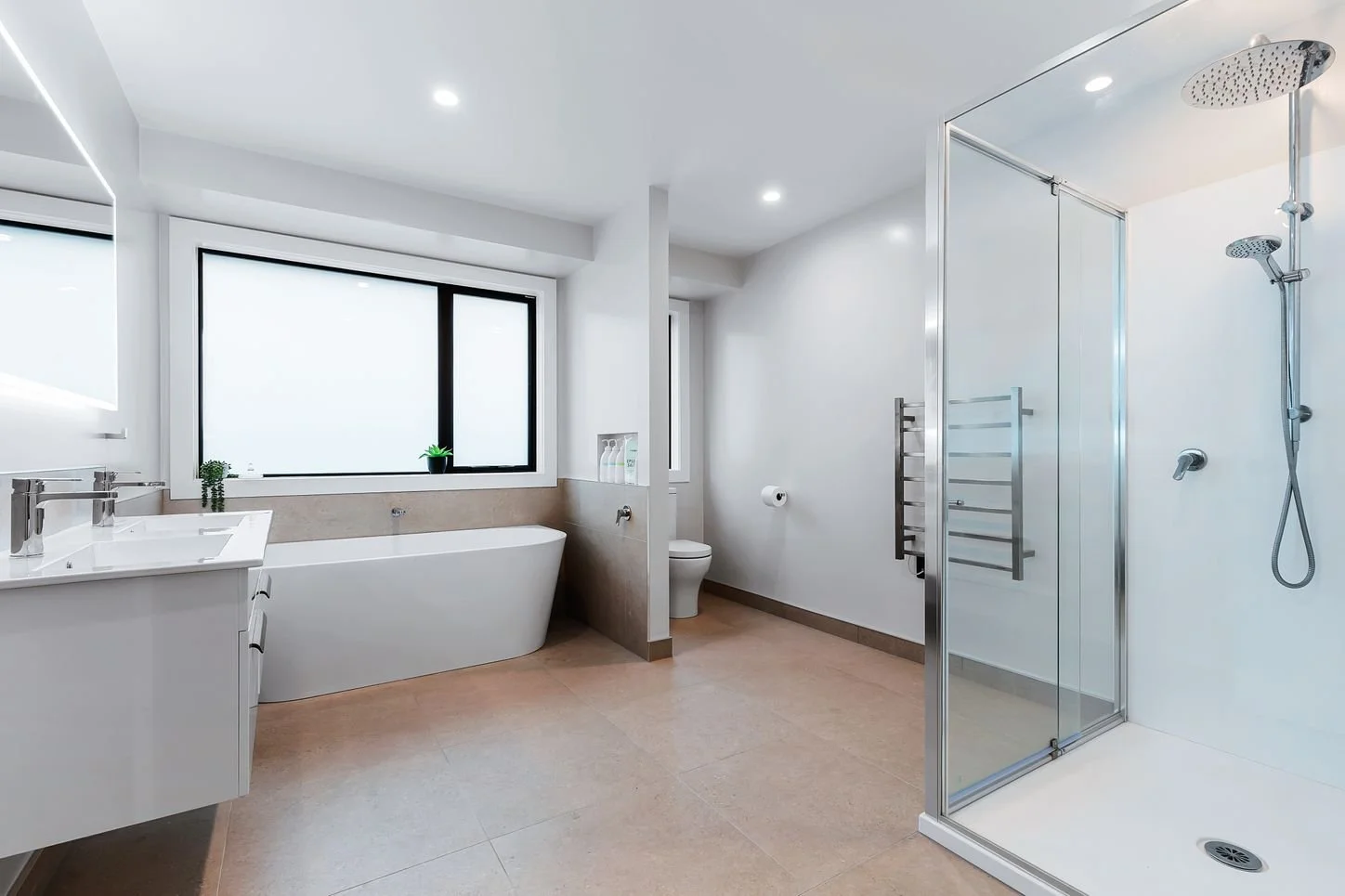 Modern bathroom with a white bathtub, large window, toilet, glass-enclosed shower, white walls, beige tiled floor, and minimal decor.