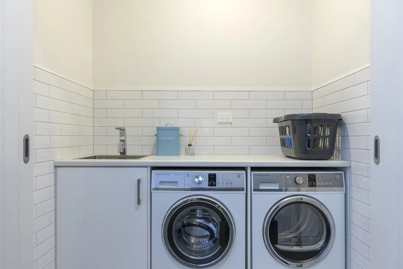 Laundry room with washer and dryer, black laundry basket on the right, small sink and countertop, wall outlet, laundry detergent box, and a reed diffuser on the countertop.