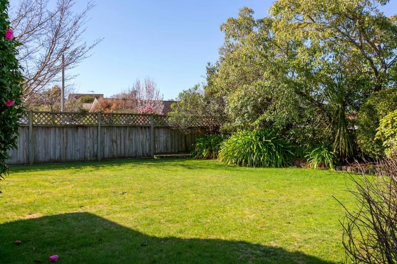 A backyard with a green lawn, wooden fence, trees, and shrubs under a clear blue sky.