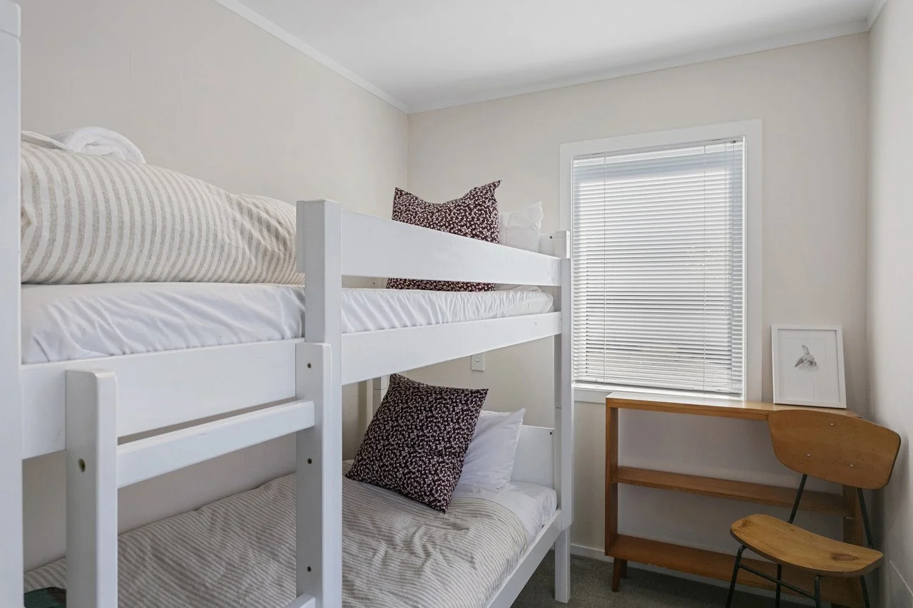 Small bedroom with white bunk beds, beige striped and white bedding, two patterned pillows, a small wooden shelf, partially closed window with blinds, framed drawing of a rabbit on shelf.