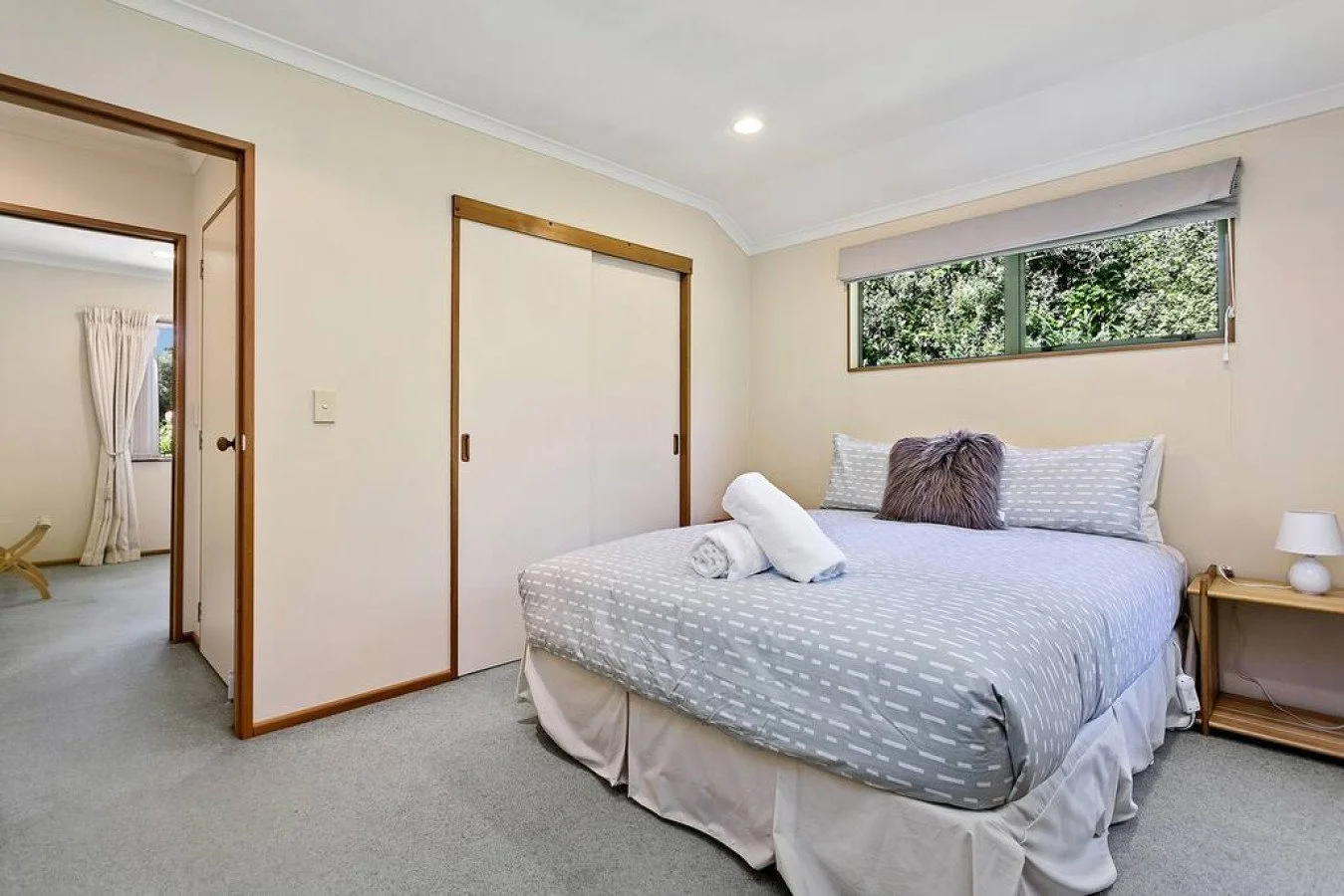 A Bedroom with a bed featuring gray and white bedding, two rolled towels, a gray pillow, and a window showing greenery outside.