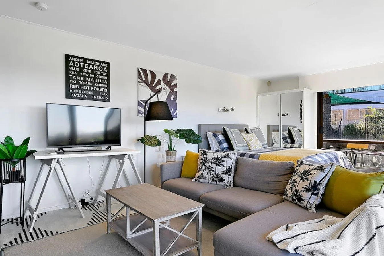 Living room with a gray couch, lion and palm tree patterned pillows, a wooden coffee table, and a TV on a white stand in front of a white wall. There are plants, a black lamp, and two pieces of wall art, with a bedroom and window with an outdoor view