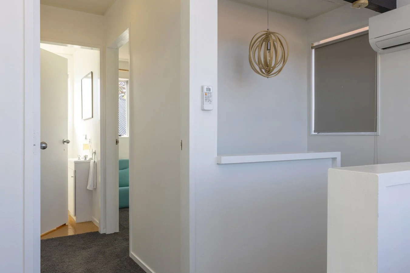 Interior of a modern home with white walls, a hanging light fixture, a window with a gray blind, an air conditioning unit, and a view into a bathroom with a small sink and a towel.