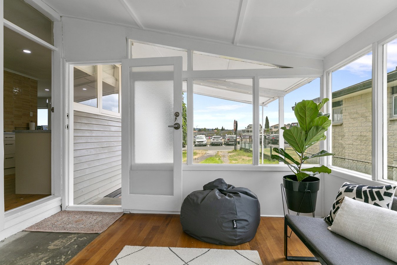 Sunroom with a white door leading outside, a large green potted plant, a gray bean bag, and a small sofa with cushions, on a hardwood floor.