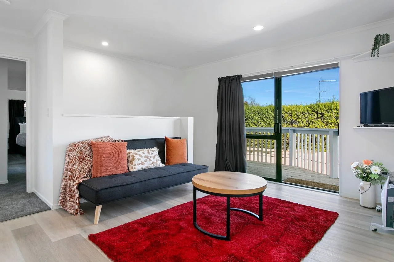 Living room with a black sofa adorned with patterned and solid throw pillows, a red area rug, a round wooden coffee table, and sliding glass door leading to a balcony with greenery outside.