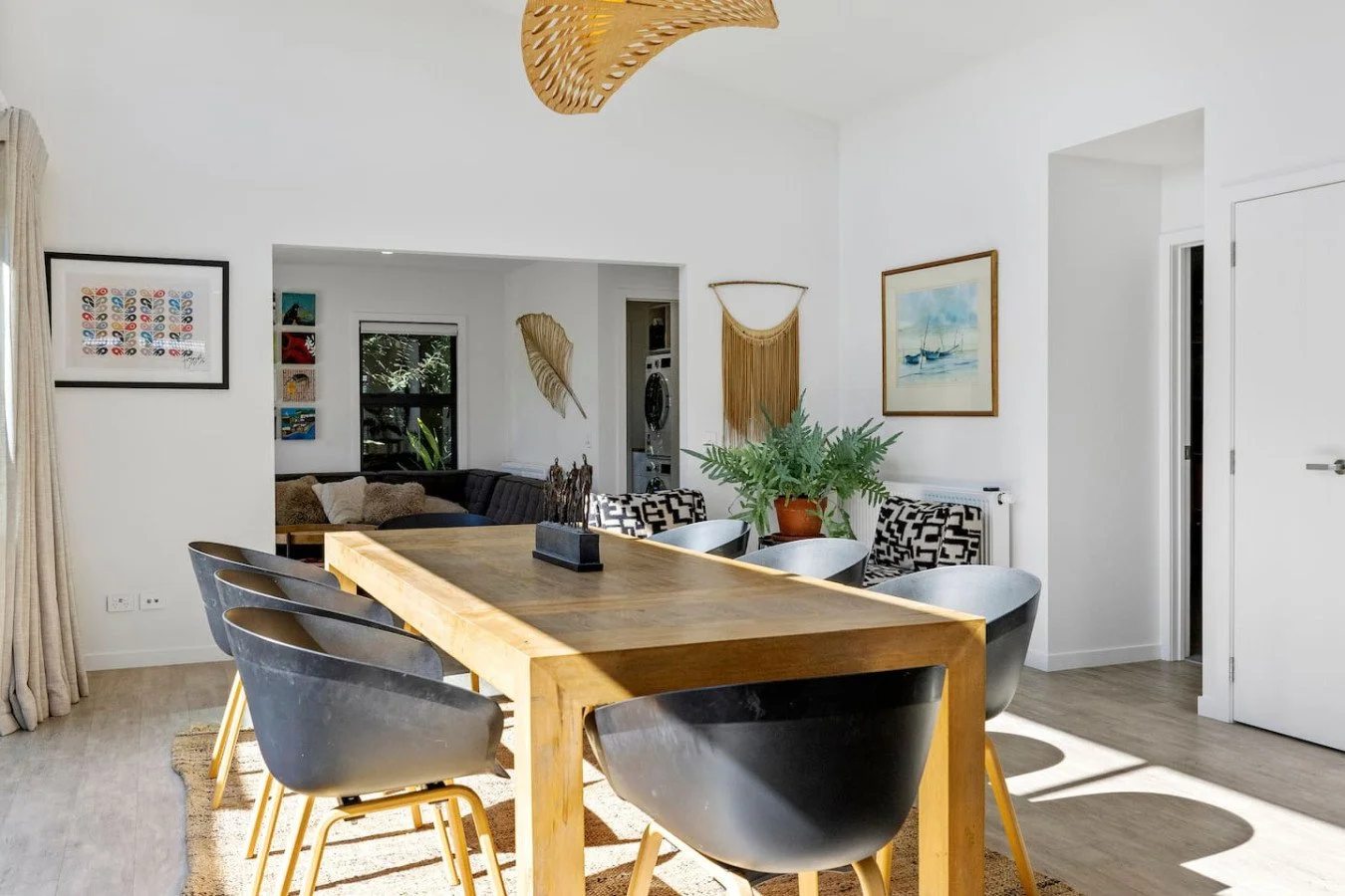 Modern dining room with a wooden table, six black chairs, a potted fern, artwork on white walls, a living area with a couch, and decor including woven wall hangings and a leaf-shaped sculpture.