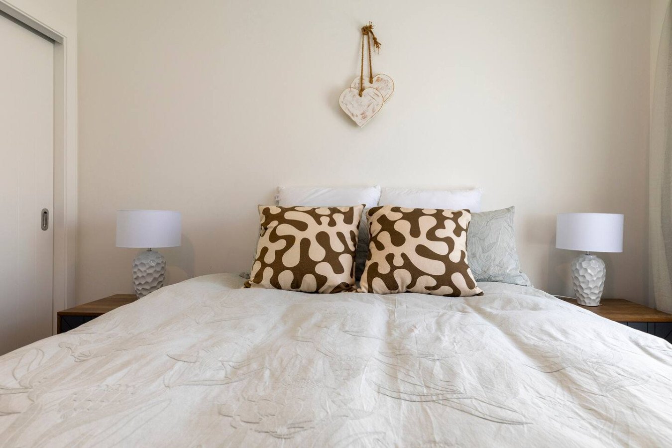 A neatly made bed with two patterned pillows, two white pillows behind, and two bedside tables with modern white lamps, against a beige wall with a heart-shaped decoration hanging above.