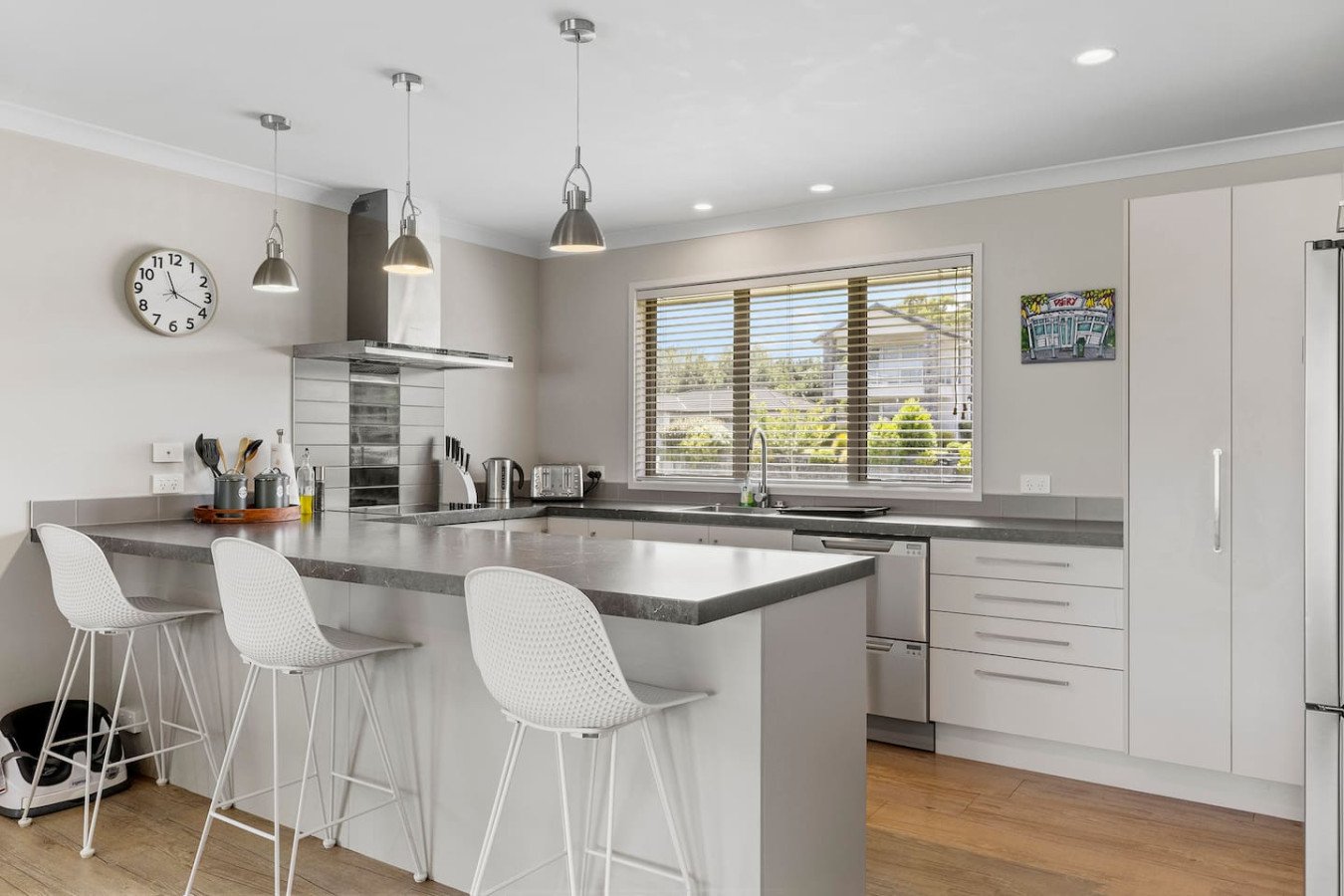 Modern kitchen with white cabinets, gray countertops, and a large window. There are three white barstools at the counter, pendant lighting, and a wall clock.