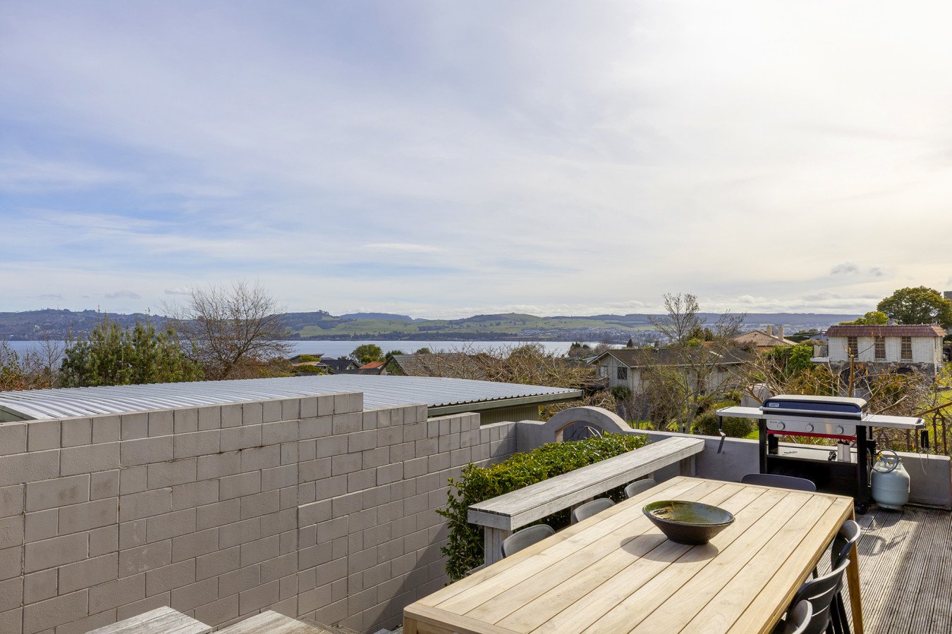 An outdoor patio with a wooden table, black chairs, and a barbecue grill. The patio overlooks a landscape of houses, trees, water, and distant hills under a partly cloudy sky.