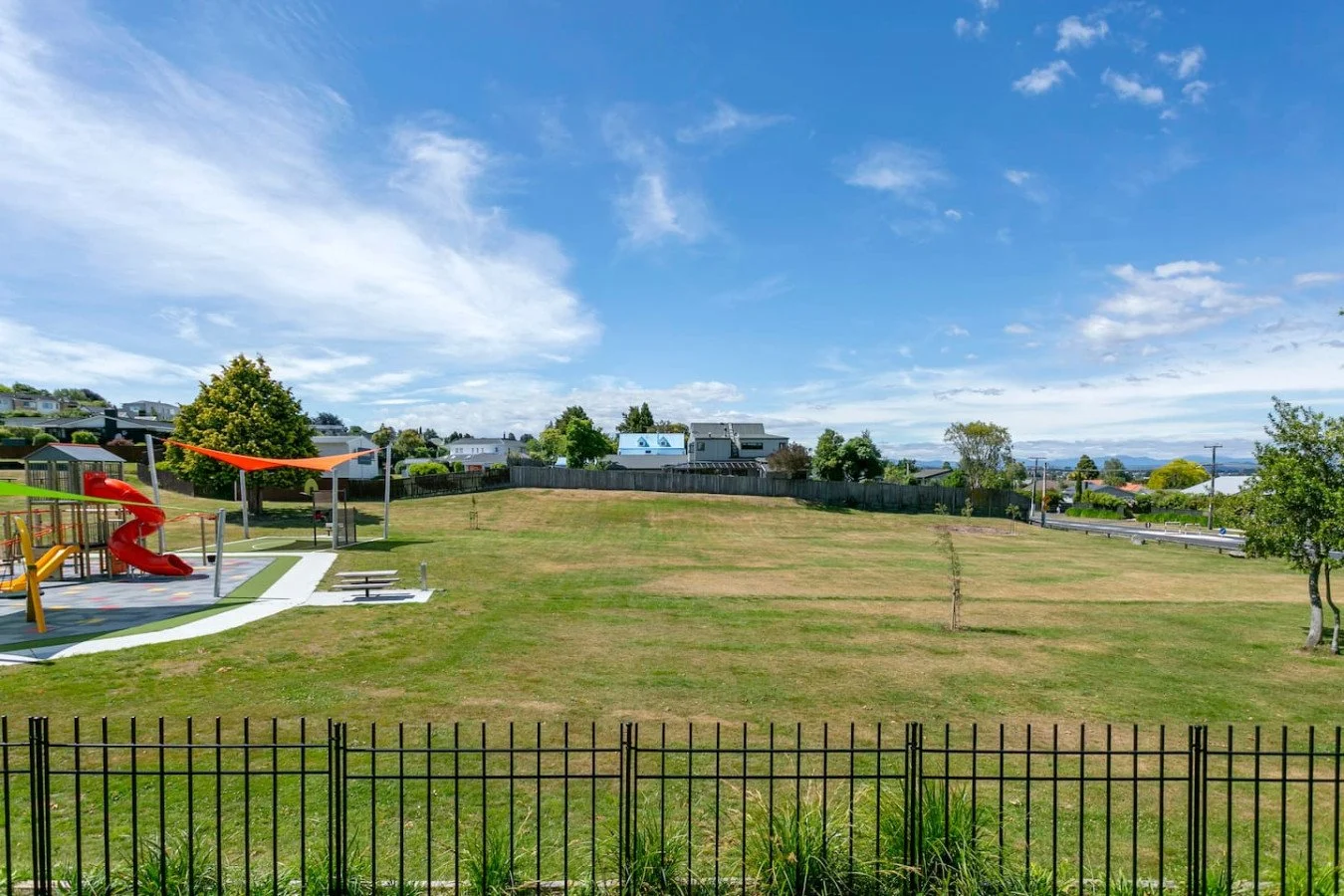 A playground with a brightly colored slide and shade sails on a grassy field under a partly cloudy sky, with trees and houses in the background.