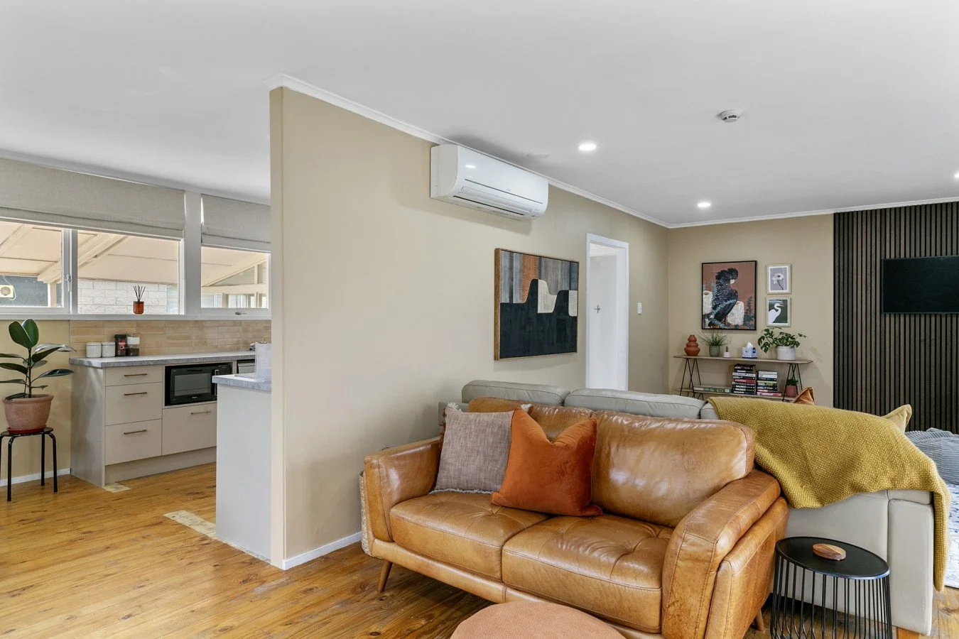 Living room with a leather couch, decorative pillows, a side table, and wall art. An air conditioning unit is mounted on the wall. Part of the kitchen with cabinetry, countertop, microwave, and a potted plant is visible in the background.