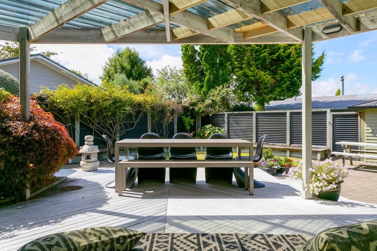 Outdoor patio with dining table and black chairs under a wooden pergola, surrounded by plants, trees, and flowers.