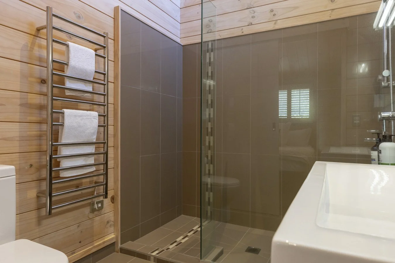 Modern bathroom with wooden and gray tiled walls, glass shower enclosure, white sink, and towel rack.