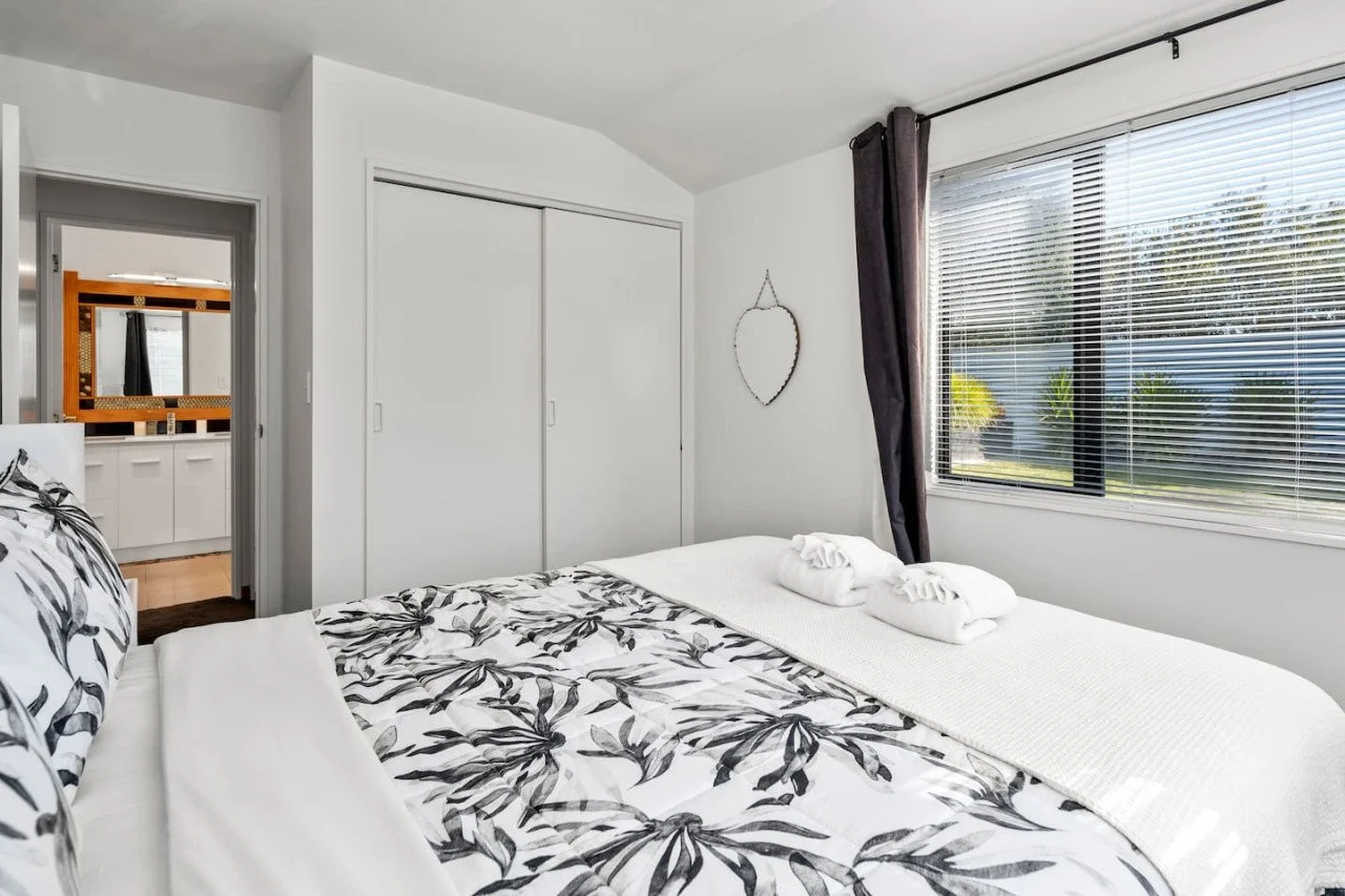 A bedroom with a large window, a bed with black and white floral bedding, and towels folded on the bed.