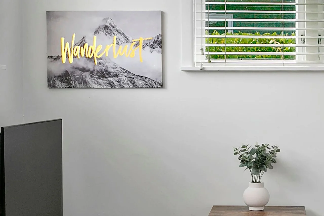 A white wall with a window with white blinds and greenery outside. A picture of snowy mountains with the word 'Wanderlust' in yellow cursive text. A wooden table with a white vase holding green leaves.
