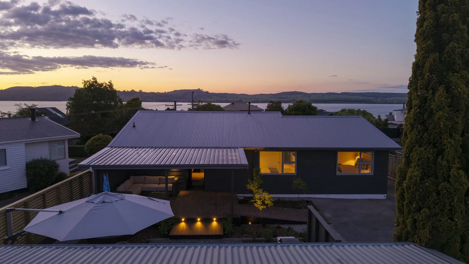 A view of a house at sunset with lit interior rooms, outdoor seating area, umbrella, and a large tree, overlooking a body of water and distant hills.