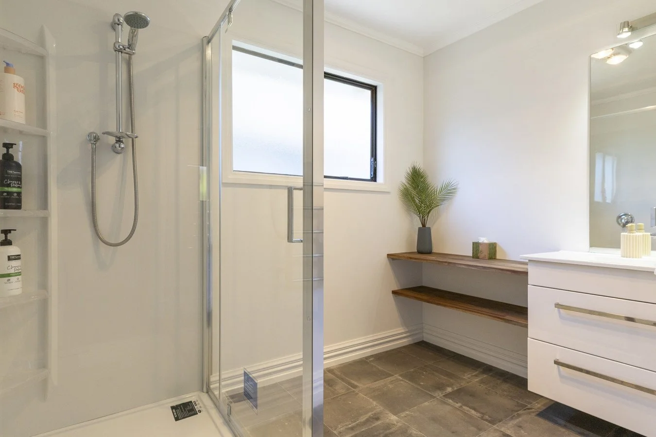 Modern bathroom with walk-in shower, white walls, a large mirror, a white vanity with drawers, a wooden shelf, a potted plant, and a frosted window.