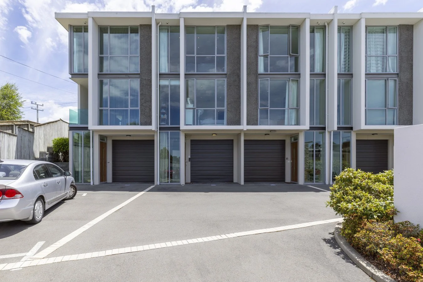 Modern multi-story residential building with four garage doors and large glass windows, parked car in front, and landscaped area on the right.
