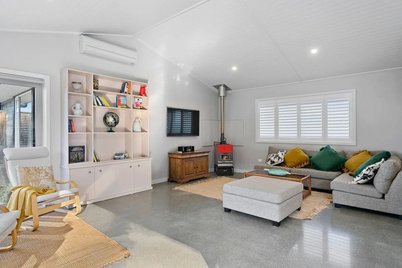 A living room with a large gray sectional sofa decorated with green and yellow pillows, a textured area rug, a flat-screen TV mounted on the wall, a small wooden coffee table, white plantation shutters on the windows, a black wood stove near the wind