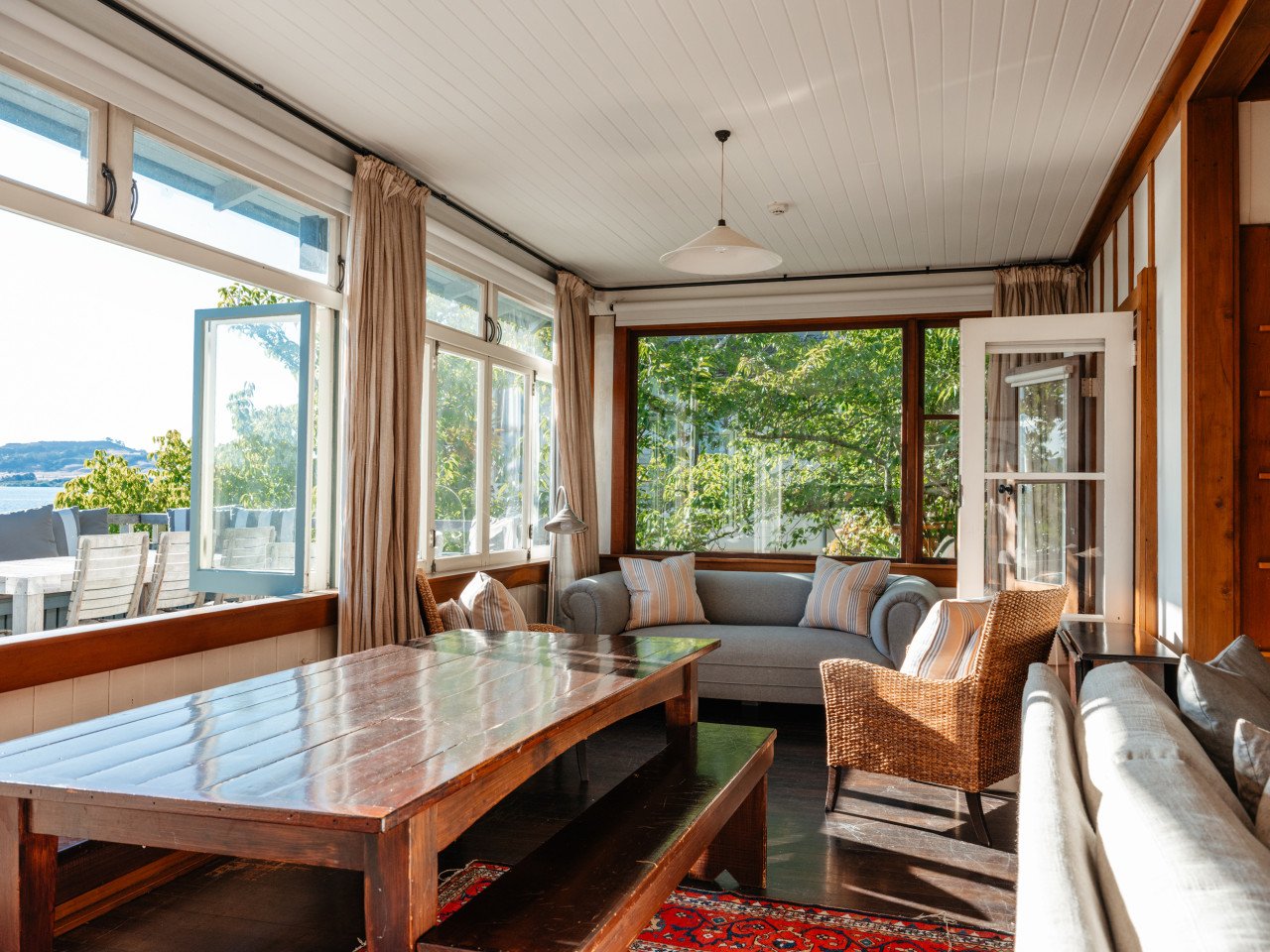 Living room with large windows showing outdoor view, a wooden dining table, a wicker chair, a gray sofa, and a small side table, decorated with striped pillows and natural light.