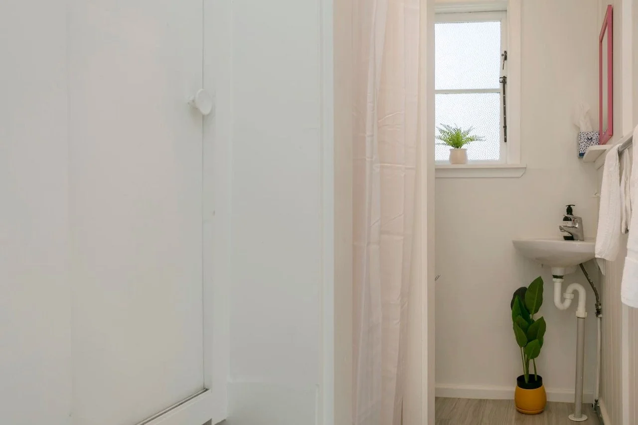 Small bathroom with a window, a potted plant on the windowsill, a wall-mounted sink with a liquid soap dispenser, a white towel hanging on a rail, a pink framed mirror, and a yellow planter with a green leafy plant on the floor.