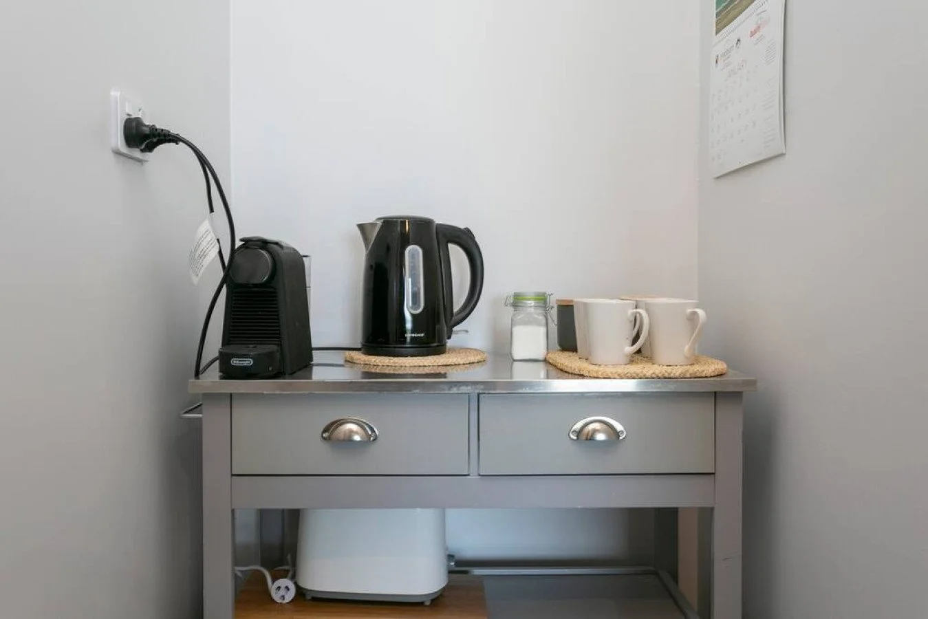 A small kitchen counter with a toaster, electric kettle, sugar container, and two mugs on woven mats; a calendar hanging on the wall.