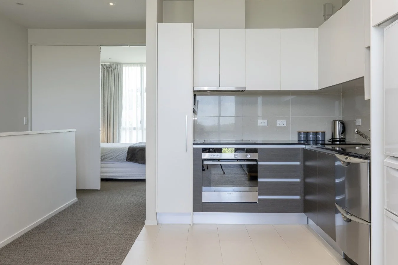 Modern kitchen with white and gray cabinets, stainless steel oven, and kitchen appliances, adjacent to a bedroom.
