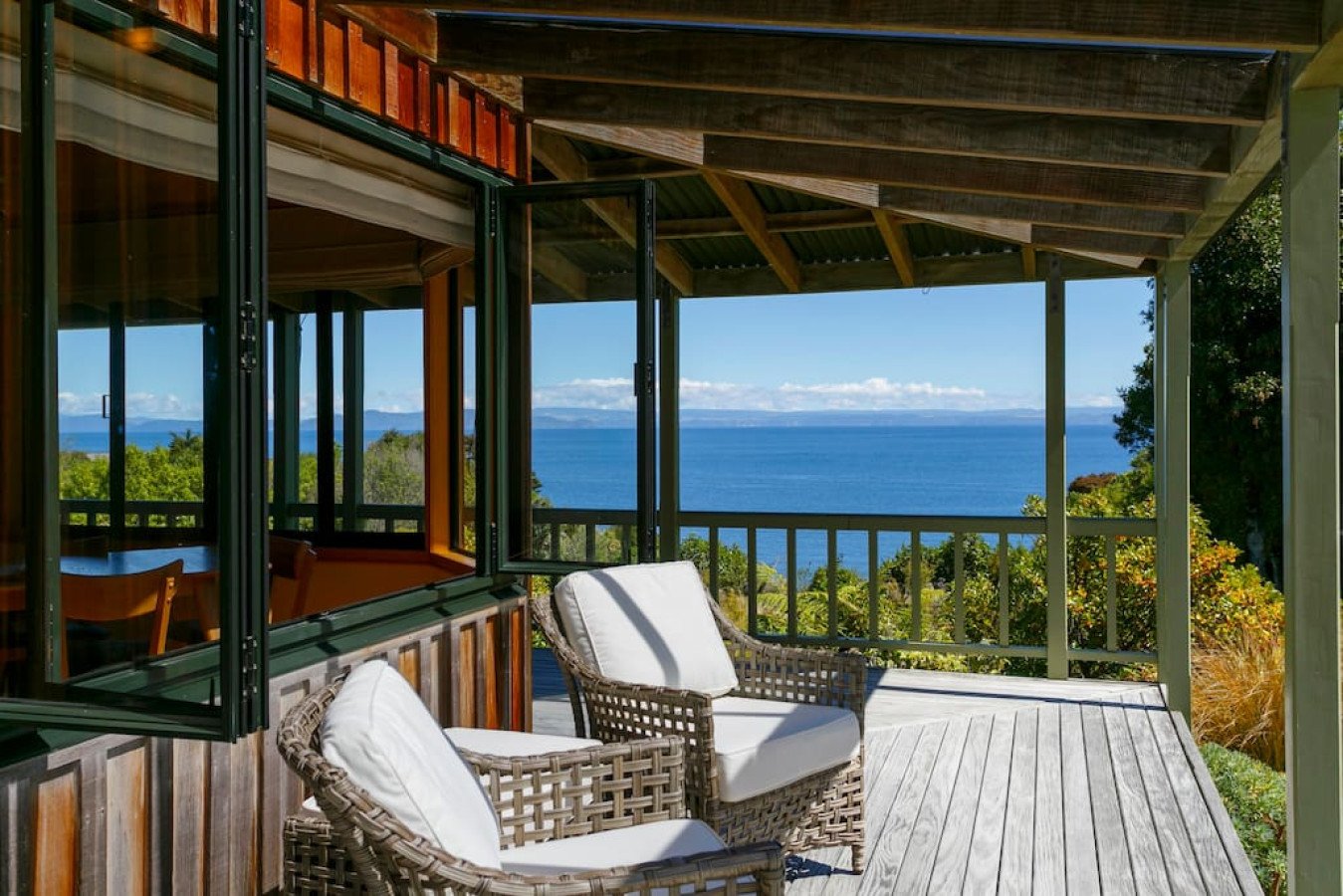 A balcony with wicker chairs and white cushions overlooking a scenic ocean view with trees and mountains in the distance.