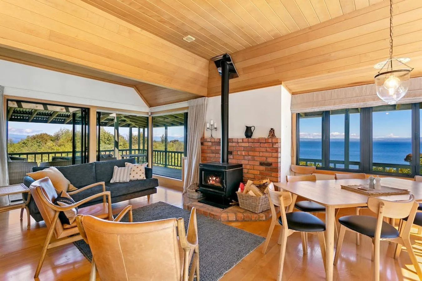 Living room with wooden ceiling, large windows showing ocean view, wood stove, brick chimney, grey sofa with pillows, wooden armchair, dining table with chairs, and a balcony outside.