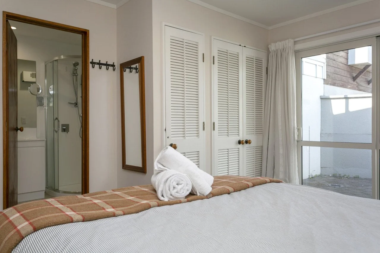 Bedroom with a bed, white towels on the bed, closet with louvered doors, window with white curtains, and a visible bathroom with a shower stall.