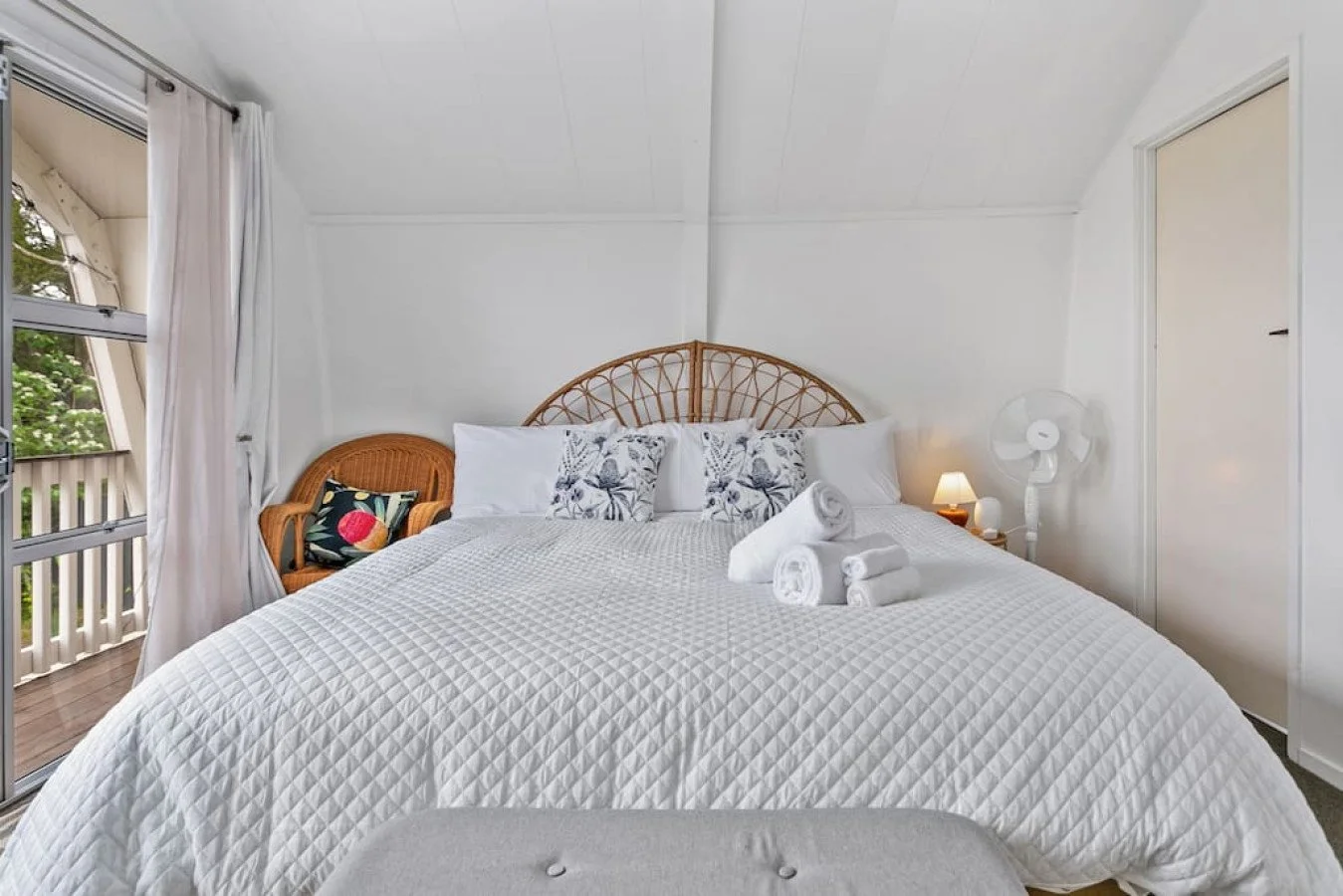 A cozy bedroom with white walls and a large bed with a white quilt, pillows, and folded towels. There is a wicker headboard, a small nightstand with a lamp and fan, a wicker chair with a decorative pillow, and a sliding glass door with white curtains