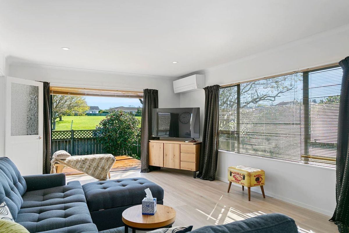 Bright living room with large windows, a black sofa, a small wooden coffee table with tissue box, a TV on a wooden stand, a small yellow ottoman with "Shell" motor oil logo, and a balcony with outdoor greenery visible through open door and window.