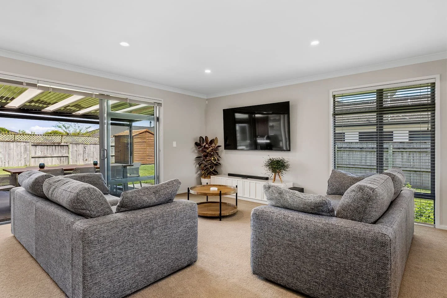 Living room with two gray sofas, a wall-mounted TV, two potted plants, a wooden coffee table, and sliding glass doors leading to an outdoor patio.