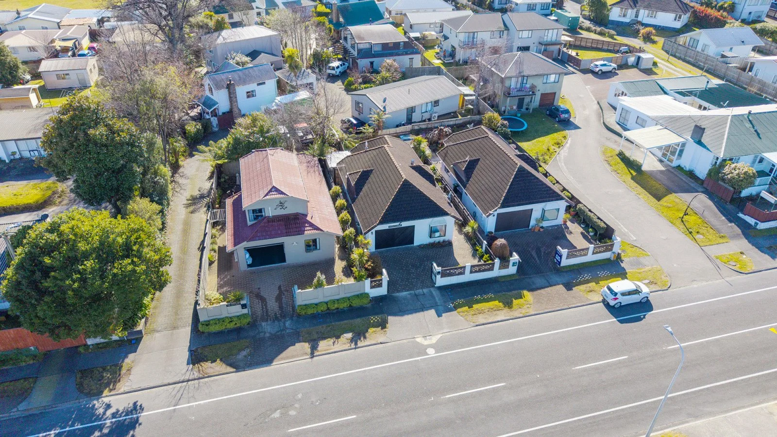 Aerial view of a neighborhood with detached houses, some with front yards and driveways, and a street with a white car parked along it. There are trees and suburban landscaping visible.