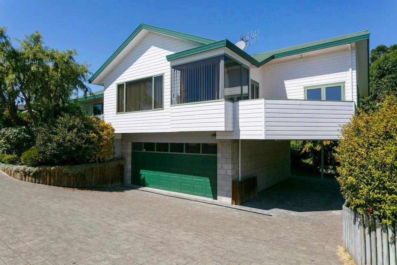 A two-story house with white siding, green roof, large windows, and a garage with a green door. The house has a balcony and is surrounded by bushes and trees with a paved driveway.