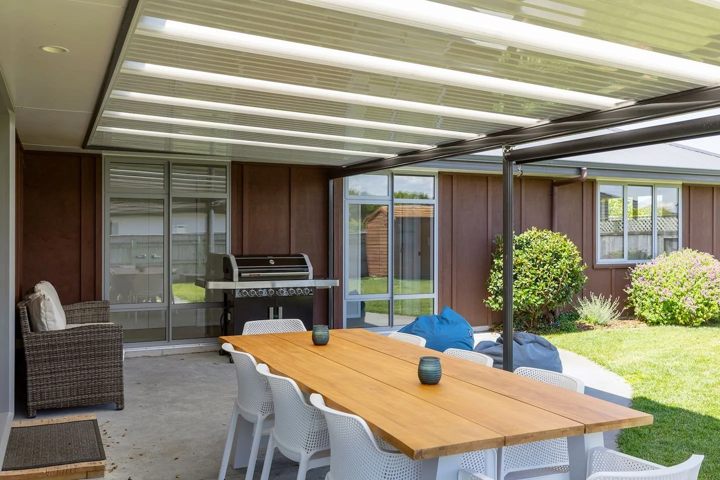 Outdoor patio with a wooden dining table, white chairs, a barbecue grill, and a wicker chair, surrounded by a garden with bushes and grass, partially covered by a pergola with clear plastic panels.