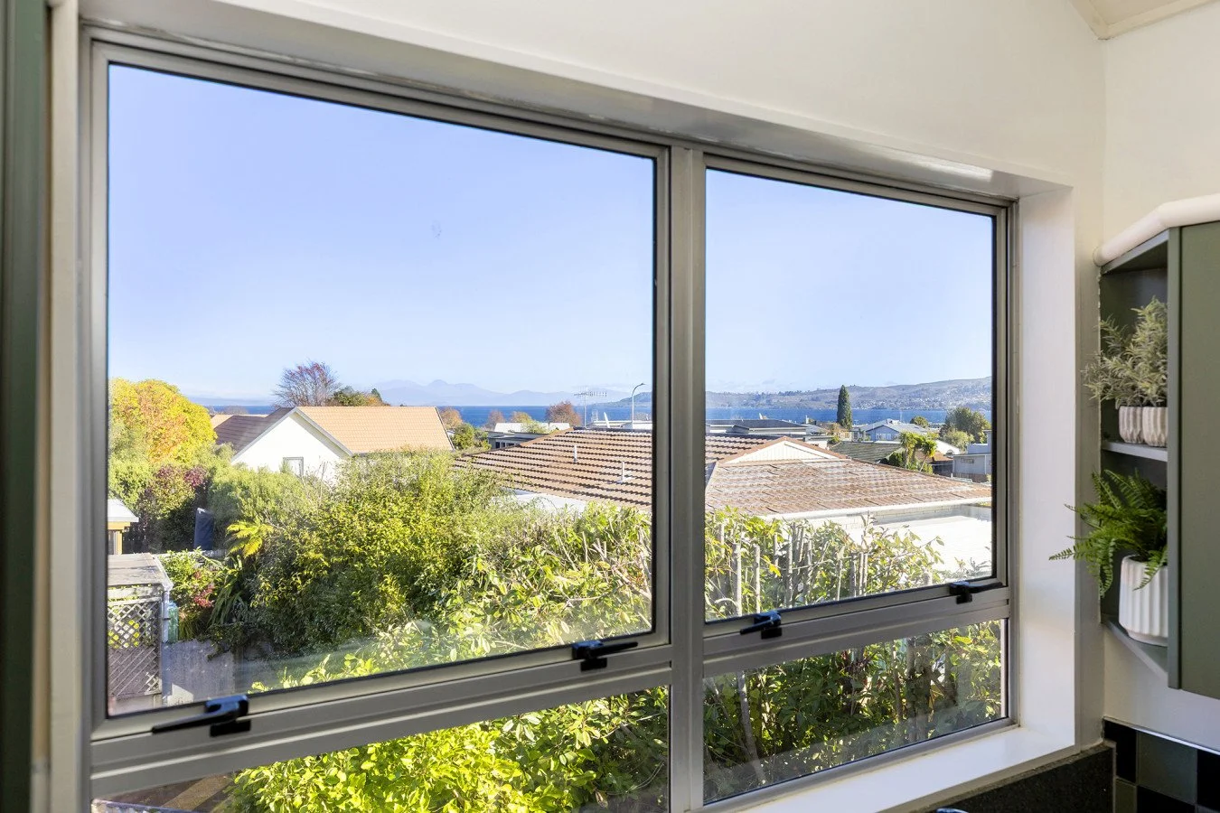 View through a kitchen window showing rooftops, trees, and distant hills and water on a clear day.
