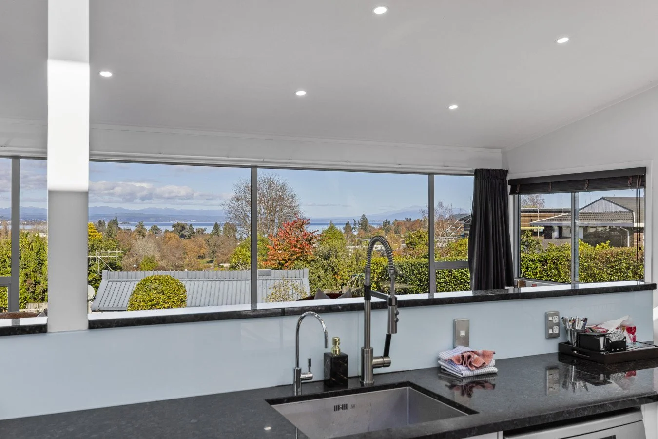 Modern kitchen with black countertop, stainless steel sink, and large windows showing a landscape with trees, houses, and a body of water in the distance.